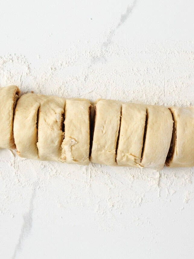 Warm, gooey, and dripping in a maple caramel glaze and pecan topping, these homemade sticky buns are the perfect bake for your next holiday brunch or cozy weekend morning! Rolled cinnamon sugar dough log on a floured surface sliced into even sections for sticky buns.