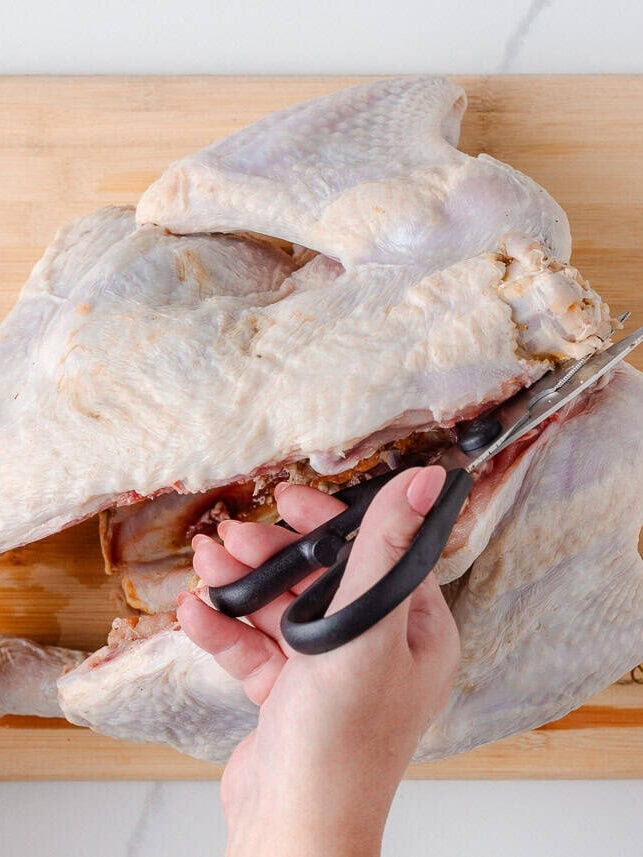 Hands using kitchen shears to cut along the backbone of a raw turkey to spatchcock it.