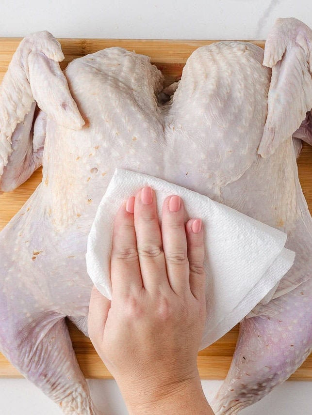 Patting a raw spatchcocked turkey dry with a paper towel on a wooden cutting board.