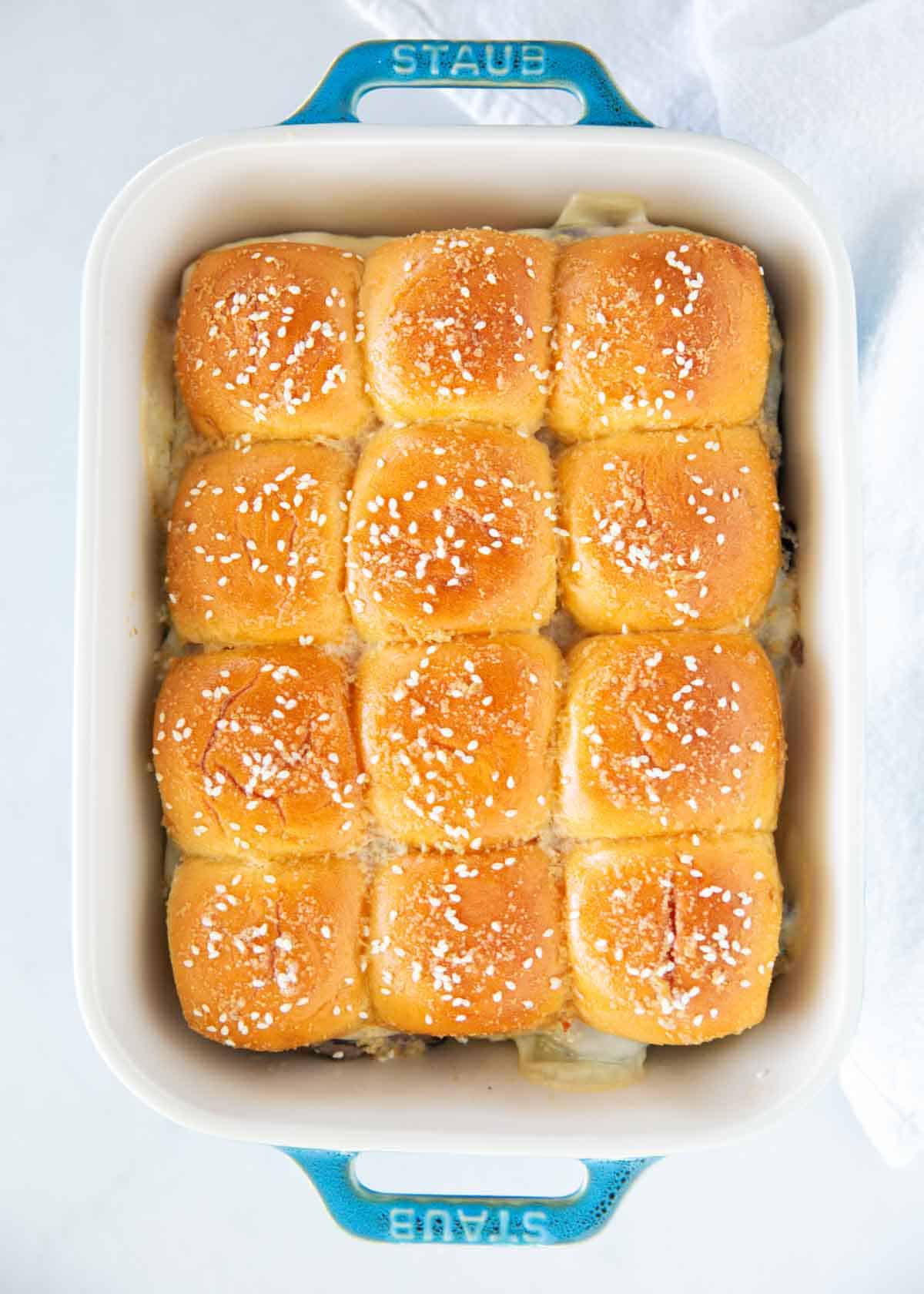 Golden baked roast beef sliders in a white Staub baking dish topped with sesame seeds.