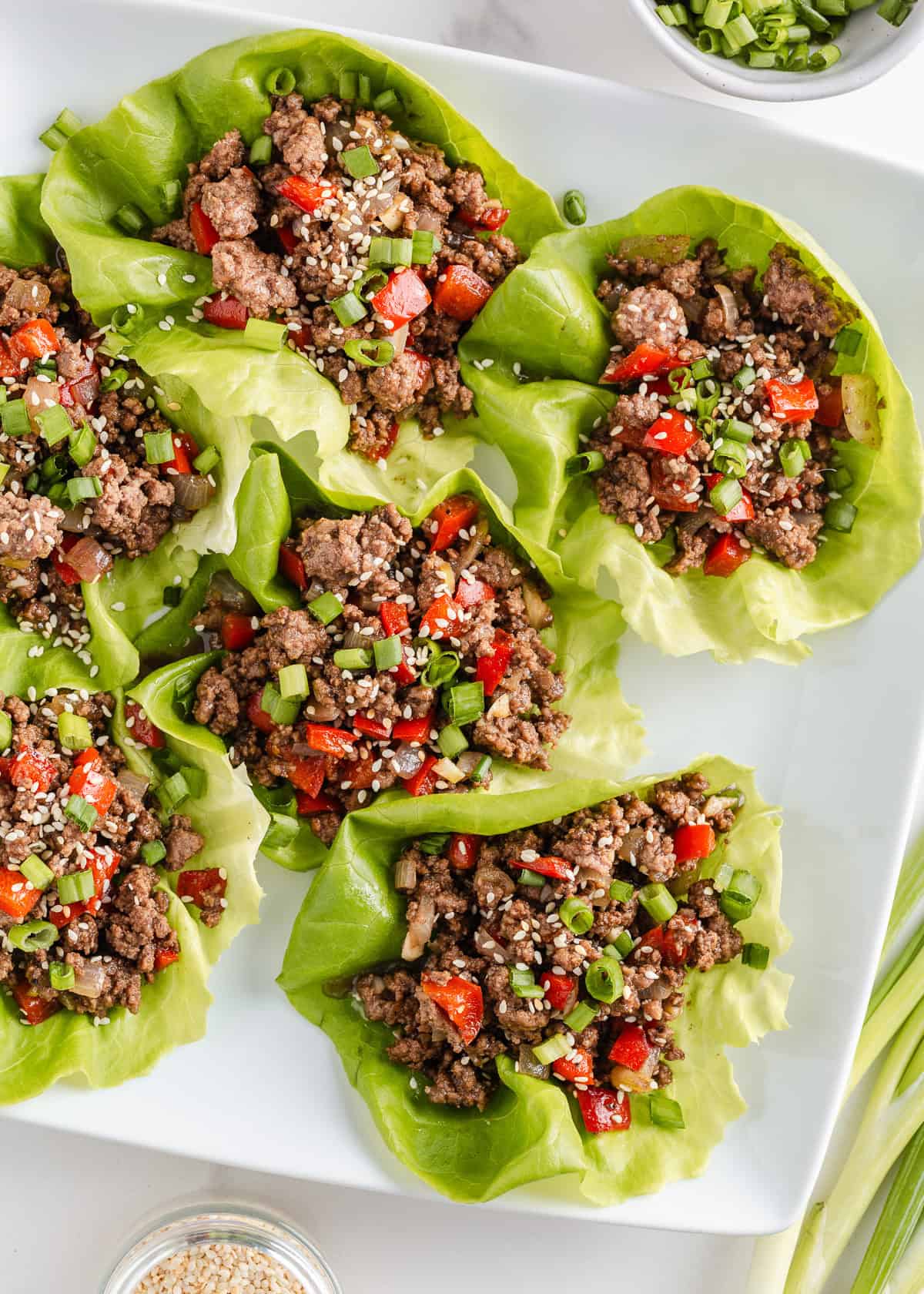 Asian-inspired ground beef lettuce wraps are easy, satisfying, and better than takeout with a juicy, sweet and spicy filling wrapped in crisp butter leaves and ready in 15 minutes. Asian-style ground beef lettuce wraps filled with seasoned beef, red bell peppers, green onions, and sesame seeds on a white platter.