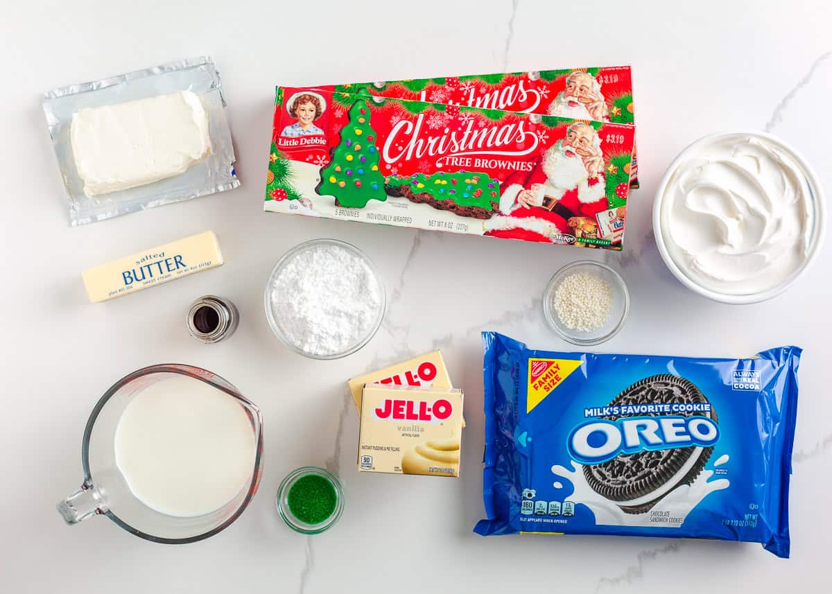 This Christmas dirt cake is a simple, no-bake dessert made with an Oreo crust, creamy whipped topping pudding layer and decorated with Little Debbie Christmas trees! Overhead view of Christmas Oreo dirt cake ingredients including Oreos, cream cheese, butter, vanilla pudding, milk, Cool Whip, sprinkles, and Little Debbie Christmas tree brownies.