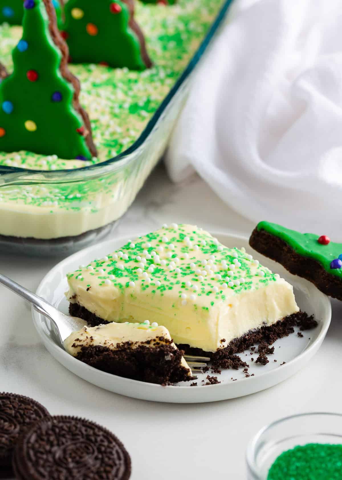 This Christmas dirt cake is a simple, no-bake dessert made with an Oreo crust, creamy whipped topping pudding layer and decorated with Little Debbie Christmas trees! Slice of Christmas dirt cake on a white plate showing creamy vanilla filling, Oreo crust, and festive sprinkles, with a Christmas tree brownie on the side.