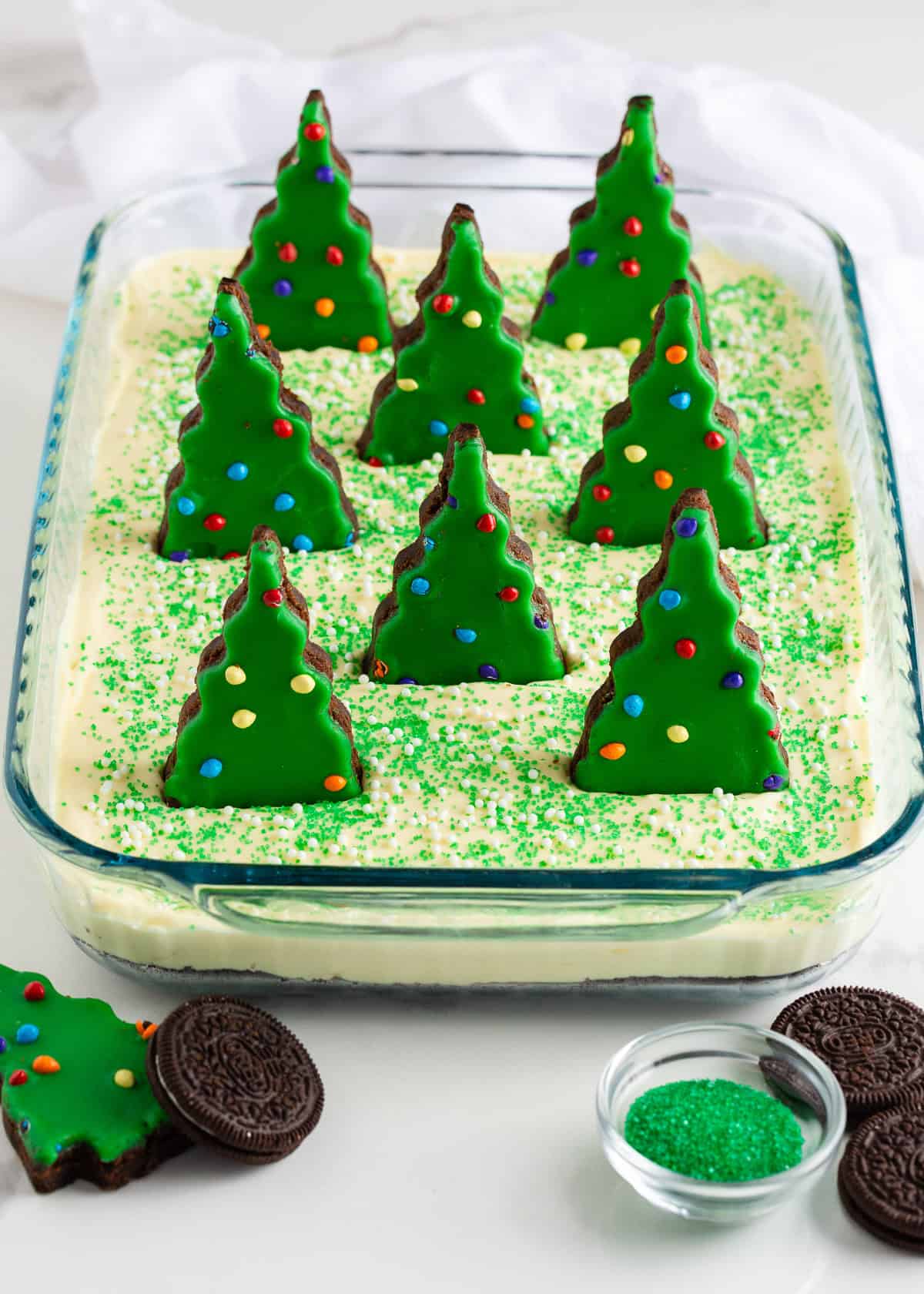 This Christmas dirt cake is a simple, no-bake dessert made with an Oreo crust, creamy whipped topping pudding layer and decorated with Little Debbie Christmas trees! Rectangular glass dish filled with Christmas dirt cake, topped with green sprinkles and Little Debbie Christmas tree brownies standing upright for a festive scene.