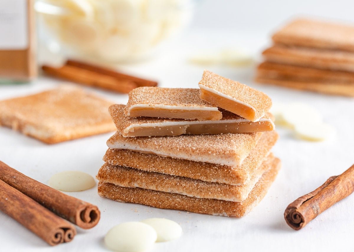 Bring home a taste of Disneyland with this churro toffee recipe. Buttery toffee coated in white chocolate and dusted with cinnamon sugar has classic churro flavor in every bite. Churro toffee squares stacked neatly with a bowl of white chocolate melting wafers behind them.