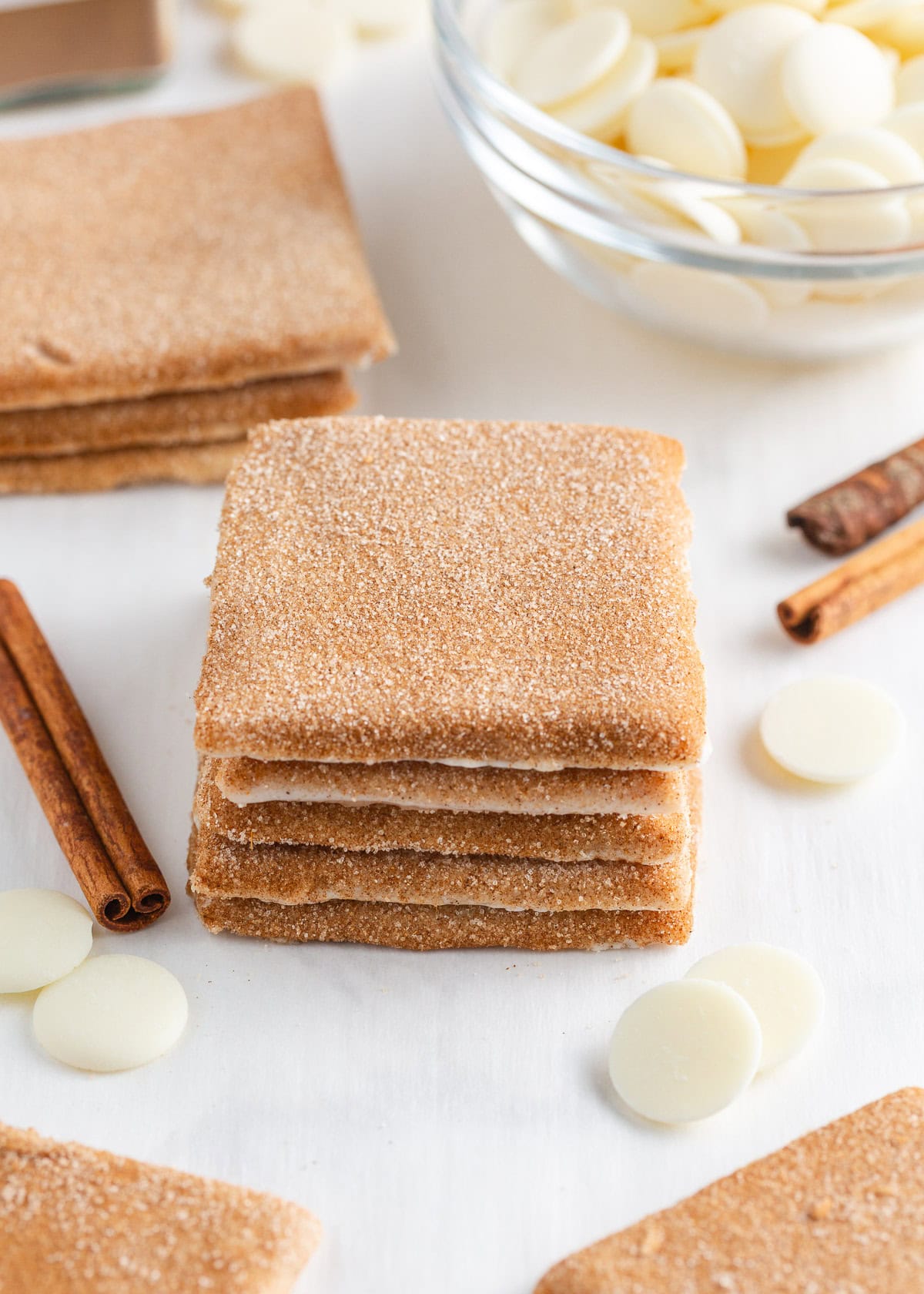 Bring home a taste of Disneyland with this churro toffee recipe. Buttery toffee coated in white chocolate and dusted with cinnamon sugar has classic churro flavor in every bite. Stack of churro toffee squares coated in cinnamon sugar on a white surface with cinnamon sticks and white chocolate wafers in the background.