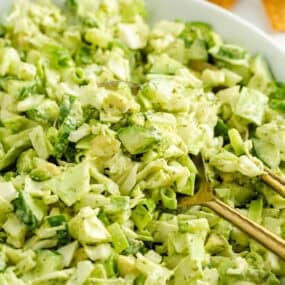 Bowl of green goddess salad with golden serving spoons and crackers in the background.