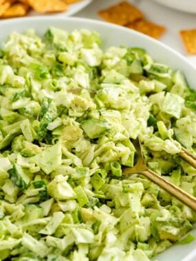 Bowl of green goddess salad with golden serving spoons and crackers in the background.