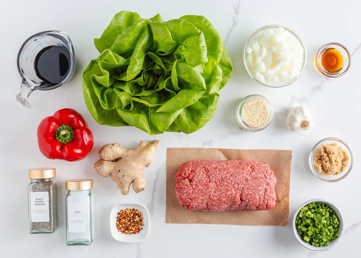 Asian-inspired ground beef lettuce wraps are easy, satisfying, and better than takeout with a juicy, sweet and spicy filling wrapped in crisp butter leaves and ready in 15 minutes. Ingredients for ground beef lettuce wraps including butter lettuce, ground beef, red bell pepper, onion, ginger, garlic, soy sauce, sesame seeds, green onions, and seasonings arranged on a white counter.