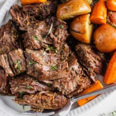 Instant Pot pot roast piled on a white platter with fork-tender carrots and baby potatoes, garnished with fresh herbs.