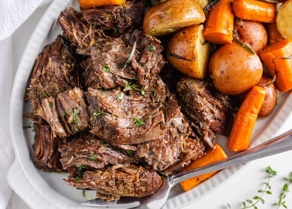 Instant Pot pot roast piled on a white platter with fork-tender carrots and baby potatoes, garnished with fresh herbs.
