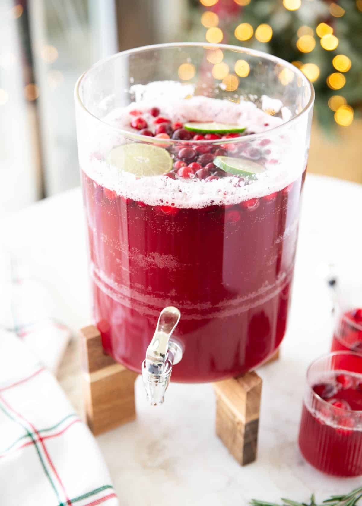 This easy cranberry punch is fizzy, festive, and perfect for holiday gatherings. Just mix juices, add fruit on top, and you’ve got a crowd-pleasing Christmas drink everyone loves. Clear drink dispenser filled with bright red cranberry punch, garnished with cranberries and lime slices, sitting on a wooden stand.