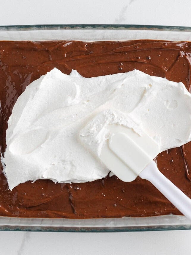 Spreading a layer of whipped topping over the chocolate pudding in a glass baking dish.
