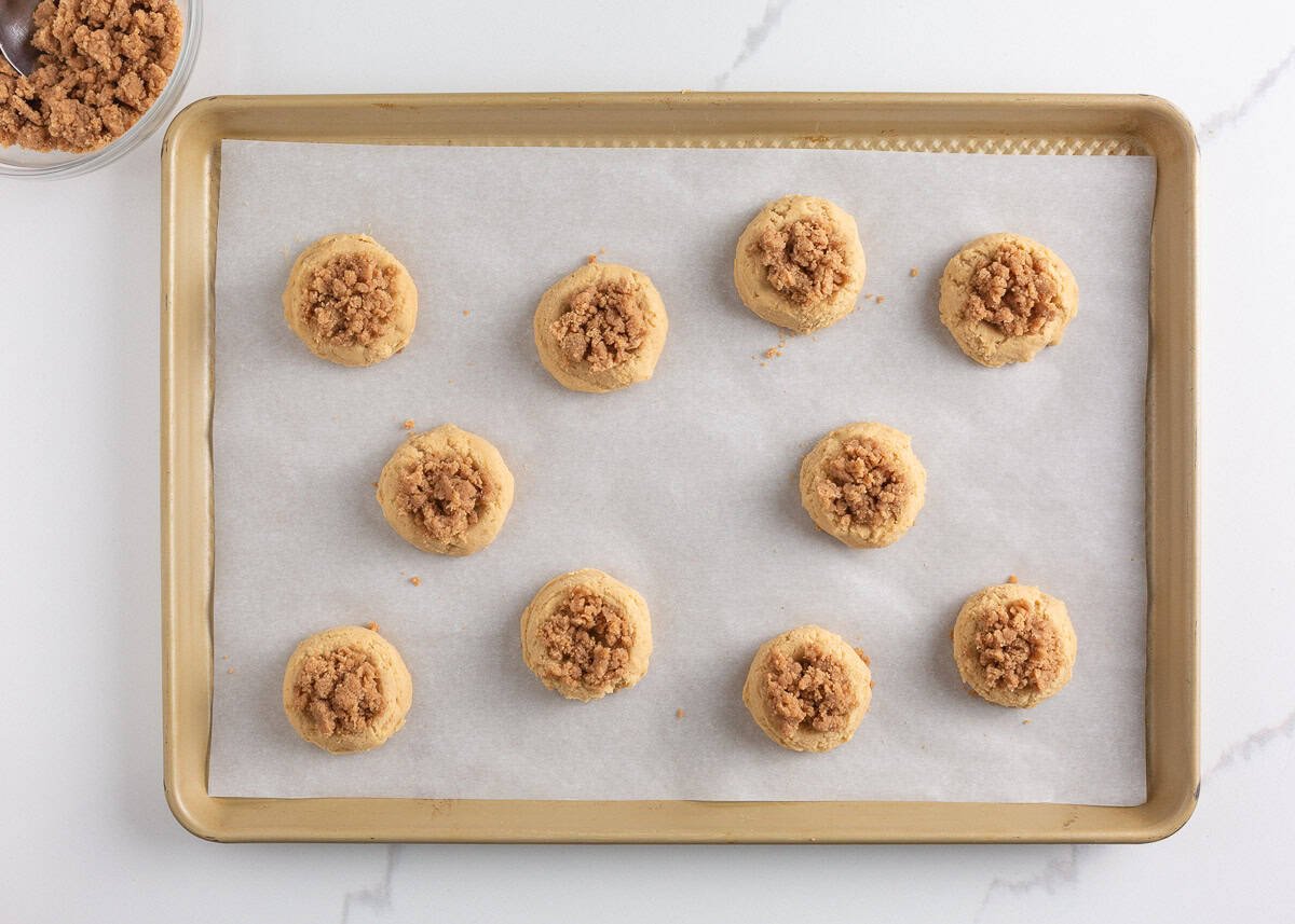 Originally inspired by The Gilmore Girls, these coffee cake cookies are made with a soft and chewy cinnamon-infused brown sugar cookie with a crumb topping and vanilla glaze! Unbaked coffee cake cookie dough balls on a parchment-lined baking sheet with cinnamon crumb topping.
