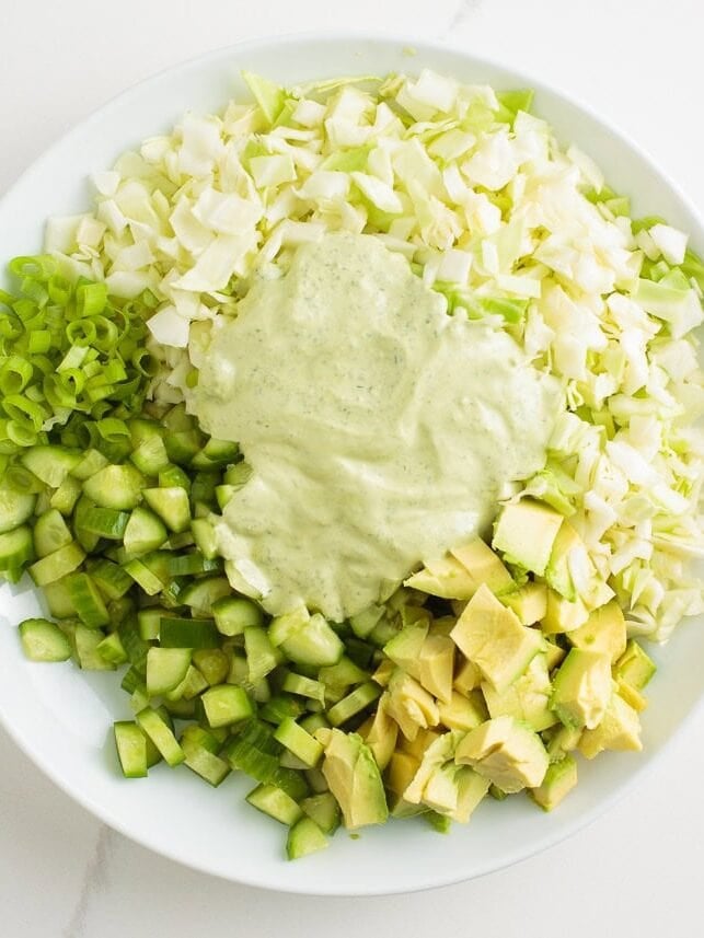 Learn to make this TikTok Green Goddess Salad with the best, most flavorful homemade green goddess salad dressing you're going to want to put on everything! Creamy green goddess dressing poured over chopped cabbage, cucumbers, avocado, and green onions in a mixing bowl.