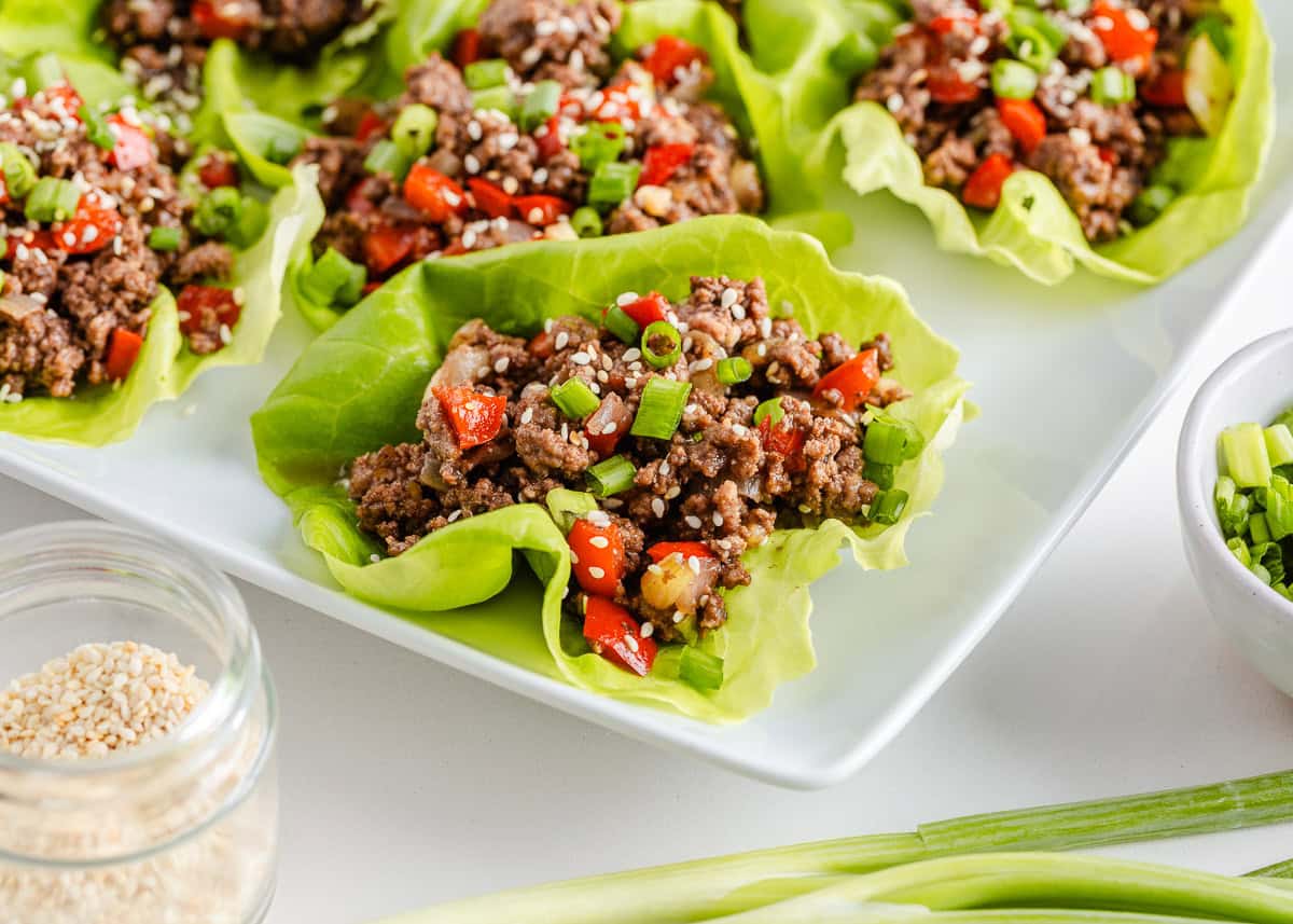 Asian-inspired ground beef lettuce wraps are easy, satisfying, and better than takeout with a juicy, sweet and spicy filling wrapped in crisp butter leaves and ready in 15 minutes. Ground beef lettuce wraps filled with seasoned beef, red peppers, green onions, and sesame seeds on a white platter.