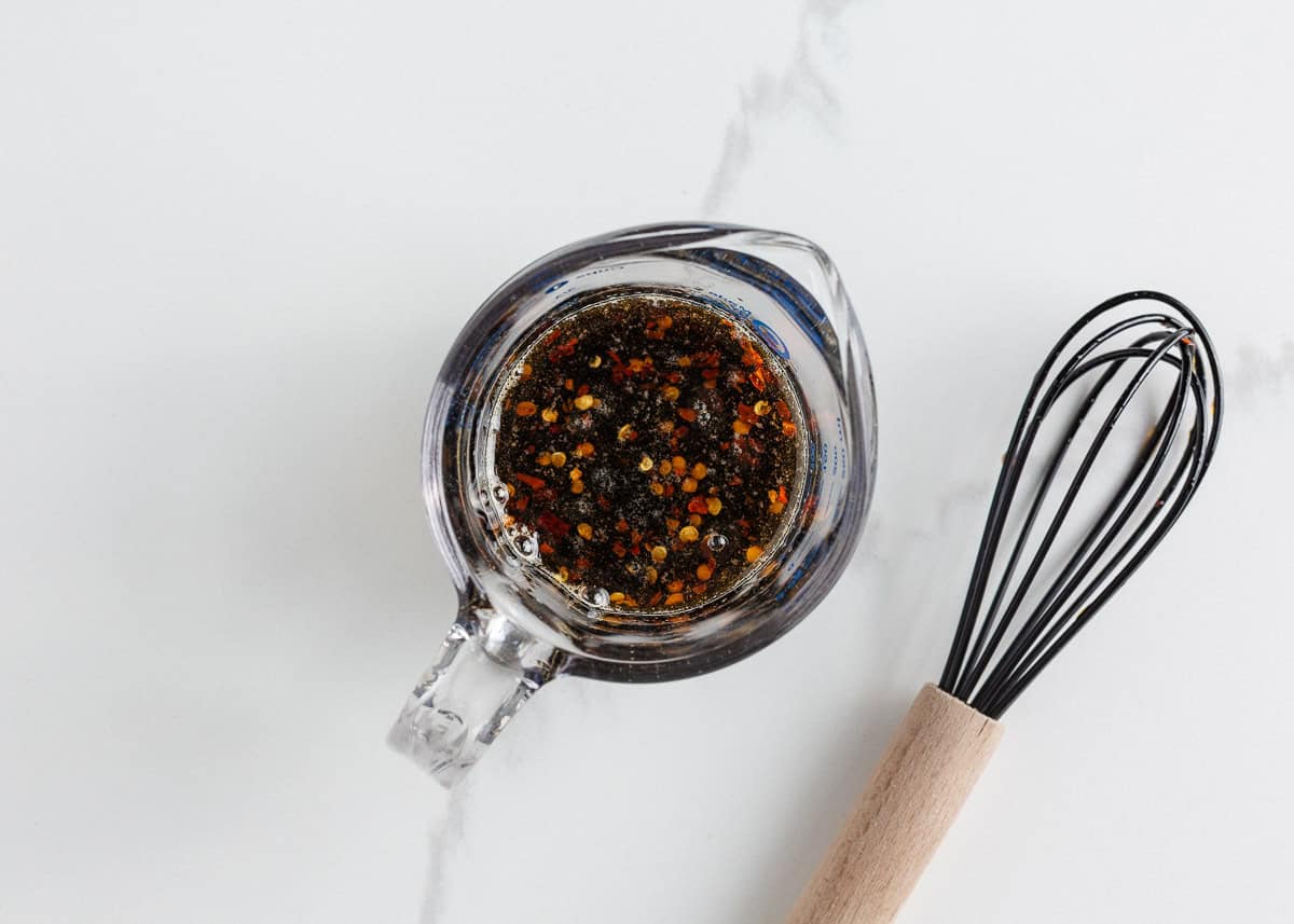 Asian-inspired ground beef lettuce wraps are easy, satisfying, and better than takeout with a juicy, sweet and spicy filling wrapped in crisp butter leaves and ready in 15 minutes. Asian-style sauce for lettuce wraps in a glass measuring cup with red pepper flakes, soy sauce, and brown sugar next to a whisk.