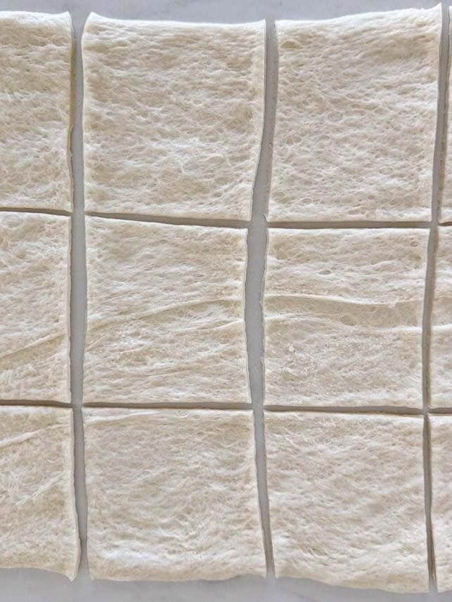 Refrigerated pizza dough rolled out and sliced into 12 even squares on a white surface.