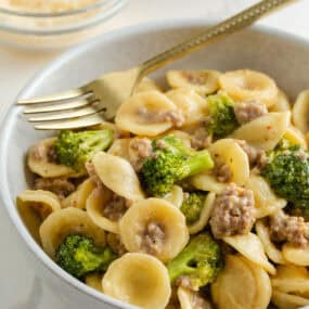 A close-up bowl of orecchiette pasta mixed with sausage and broccoli, with a gold fork resting in the bowl.