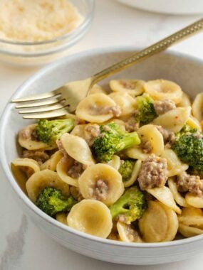 A close-up bowl of orecchiette pasta mixed with sausage and broccoli, with a gold fork resting in the bowl.