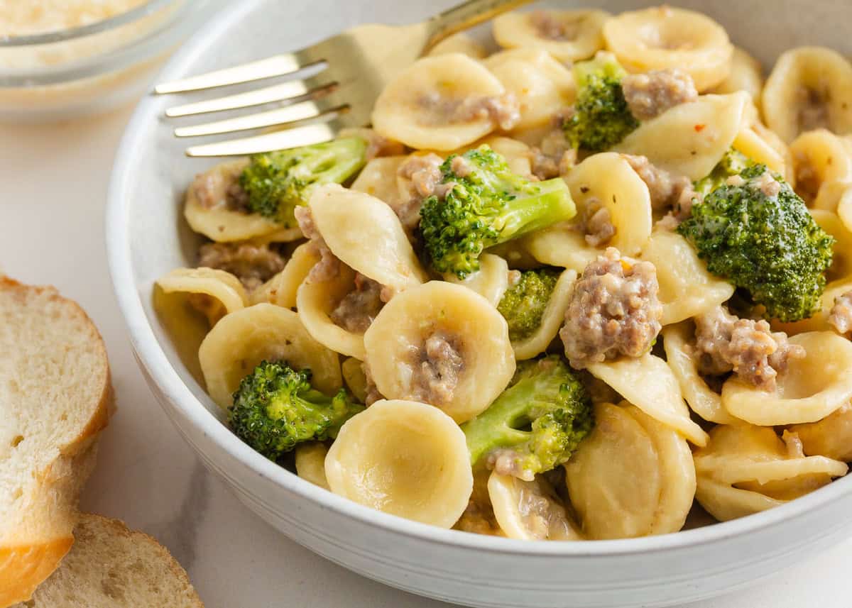 A bowl of orecchiette pasta coated in a creamy sauce with pieces of sausage and broccoli, served with slices of bread on the side.
