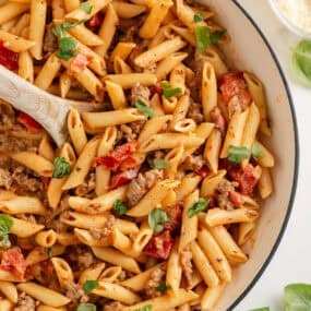 Overhead close-up of creamy Italian sausage penne pasta in a white skillet, mixed with sausage and tomatoes and topped with fresh basil.