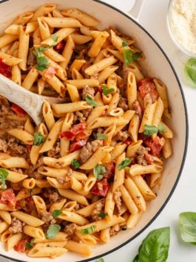 Overhead close-up of creamy Italian sausage penne pasta in a white skillet, mixed with sausage and tomatoes and topped with fresh basil.