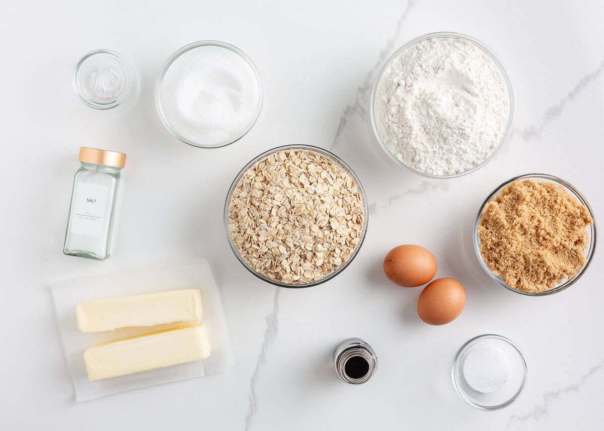 These easy oatmeal cookies are a quick treat made in only one bowl. They turn perfectly every time with a buttery brown sugar taste, crisp edges and a soft, chewy center! Overhead view of oatmeal cookie ingredients including oats, flour, brown sugar, butter, eggs, sugar, vanilla and salt arranged on a white counter.
