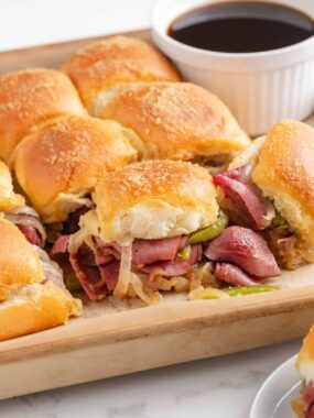Warm Philly cheesesteak sliders with toasted tops and melty provolone, packed with beef, peppers, and onions, served next to a bowl of au jus.