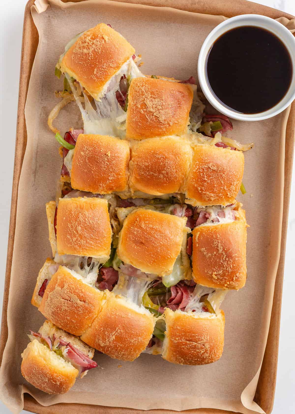 Philly cheesesteak sliders made with Hawaiian rolls stuffed with roast beef, sautéed peppers and onions, and melty cheese. A crowd-pleasing mini version of the classic! Baked Philly cheesesteak sliders on a sheet pan with melted provolone and a cup of warm au jus for dipping.