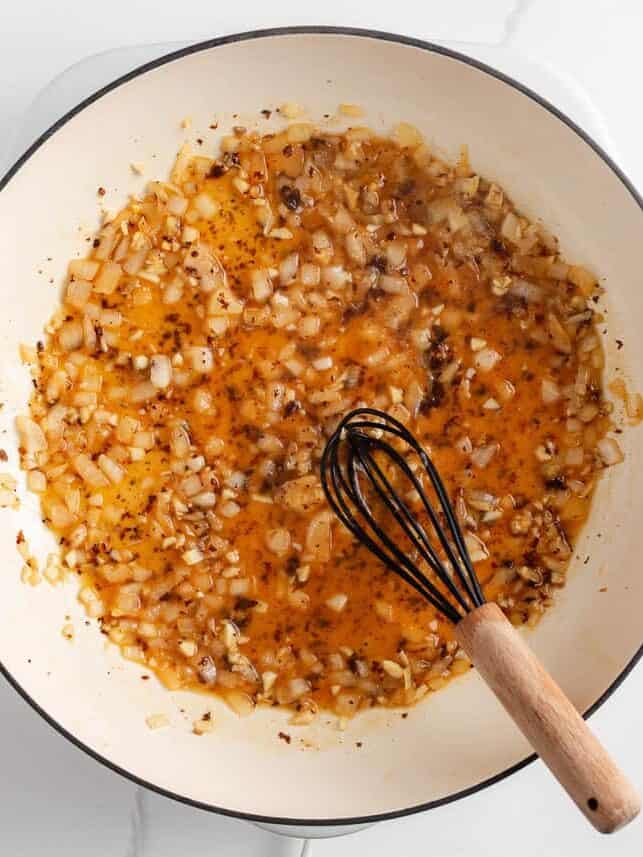 A creamy, zesty pasta recipe is something everyone needs for busy days and cozy weekends. Italian sausage pasta is a family favorite and easy to make in 30 minutes! Diced onions and garlic sautéing in broth and pan drippings in a white skillet with a whisk.