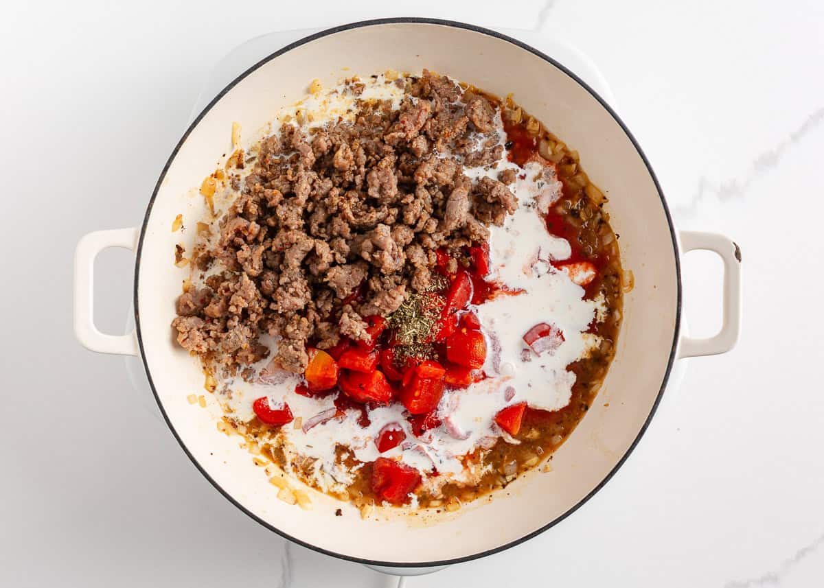 A creamy, zesty pasta recipe is something everyone needs for busy days and cozy weekends. Italian sausage pasta is a family favorite and easy to make in 30 minutes! Cooked Italian sausage, tomatoes, Italian seasoning, and cream added to sautéed onions in a white skillet before mixing.