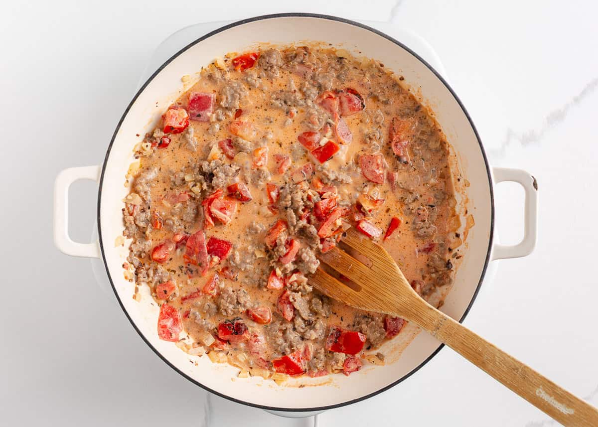 A creamy, zesty pasta recipe is something everyone needs for busy days and cozy weekends. Italian sausage pasta is a family favorite and easy to make in 30 minutes! Simmering creamy Italian sausage tomato sauce in a white skillet, stirred with a wooden spoon.