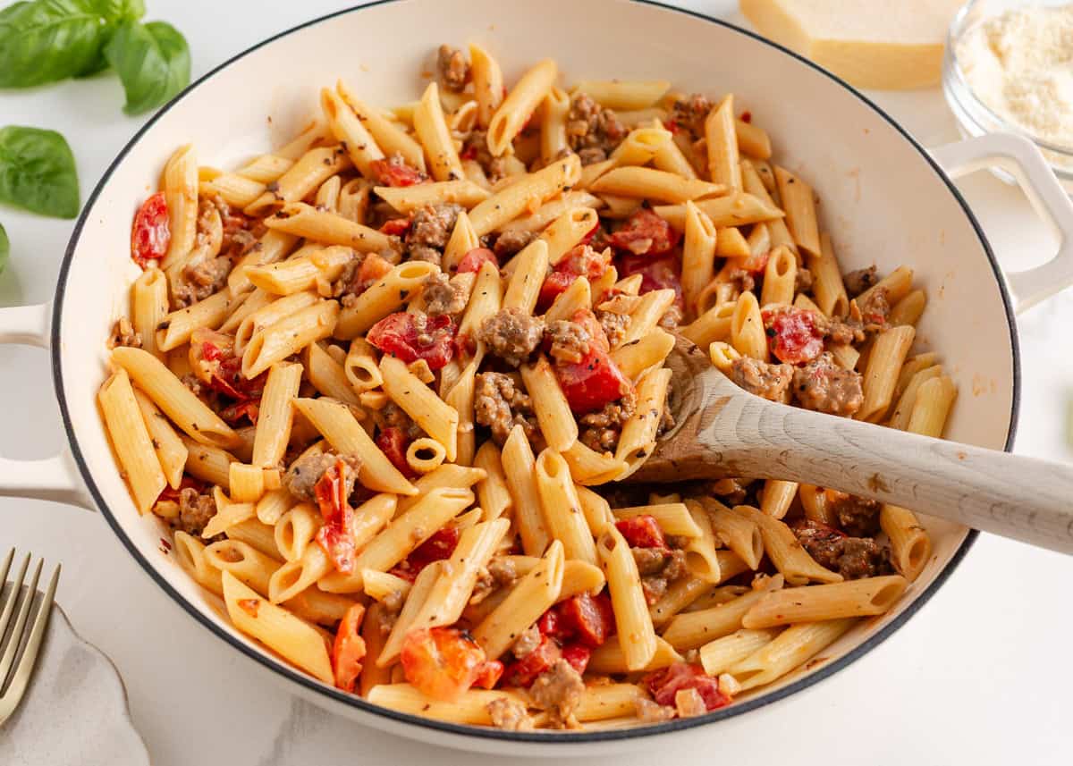 A creamy, zesty pasta recipe is something everyone needs for busy days and cozy weekends. Italian sausage pasta is a family favorite and easy to make in 30 minutes! Creamy Italian sausage penne pasta in a white skillet, mixed with sausage and tomatoes and stirred with a wooden spoon.