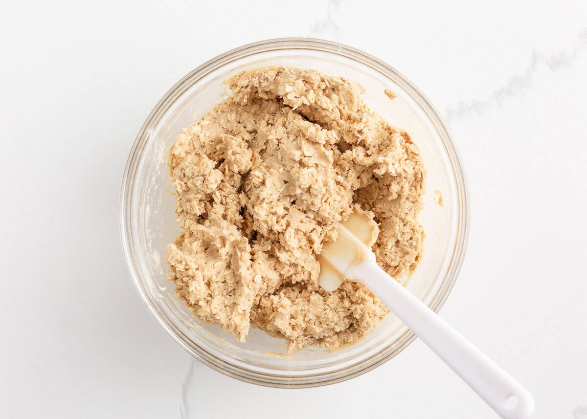 Oatmeal cookie dough mixed in a glass bowl with a spatula.