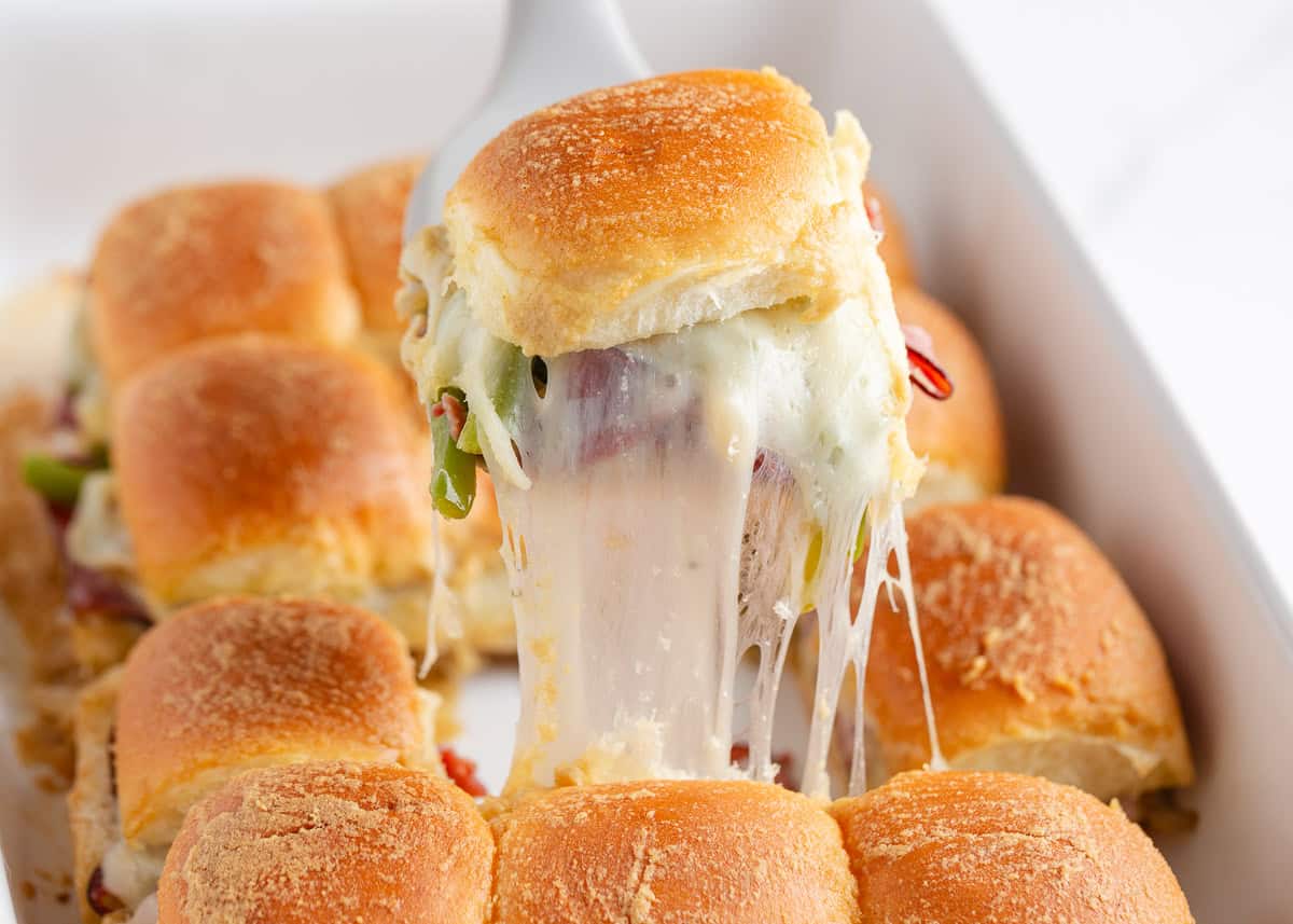 Philly cheesesteak sliders made with Hawaiian rolls stuffed with roast beef, sautéed peppers and onions, and melty cheese. A crowd-pleasing mini version of the classic! Lifting a Philly cheesesteak slider from the pan with a long, stretchy cheese pull revealing beef, peppers, and onions.