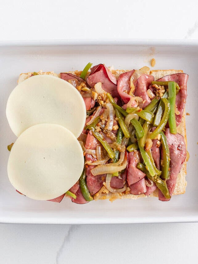 Philly cheesesteak sliders made with Hawaiian rolls stuffed with roast beef, sautéed peppers and onions, and melty cheese. A crowd-pleasing mini version of the classic! Bottom half of slider rolls topped with sautéed roast beef, peppers, onions, and sliced provolone in a baking dish.