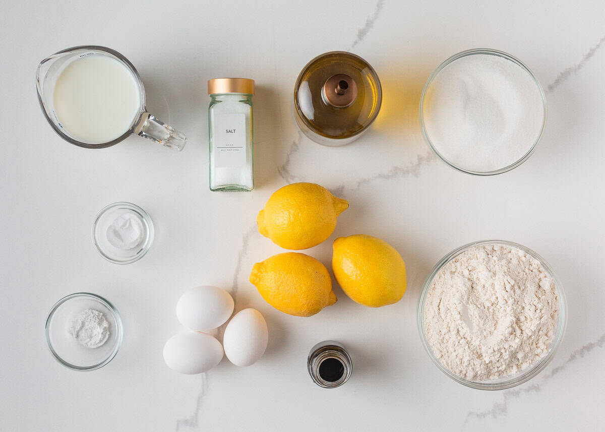 This lemon olive oil cake is sunshine in dessert form, with a light, tender crumb and bright lemon flavor in every bite. Elegant yet simple, it’s an effortless cake to make. Ingredients for lemon olive oil cake including fresh lemons, olive oil, eggs, milk, sugar, and flour on a white surface.