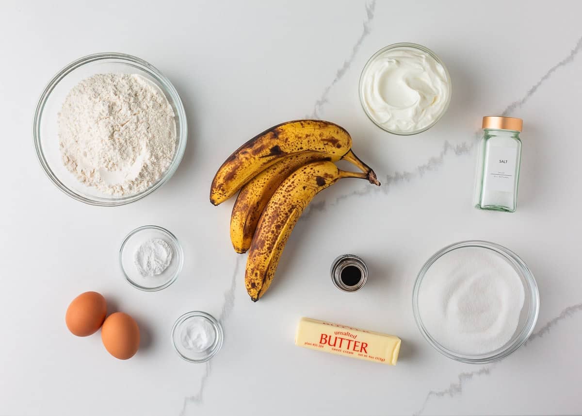 This sour cream banana bread is extra soft, moist, and full of flavor thanks to one simple ingredient. It’s an easy recipe and the perfect way to use up overripe bananas. Ingredients for sour cream banana bread including ripe bananas, flour, sugar, butter, eggs, sour cream, and vanilla on a white countertop.