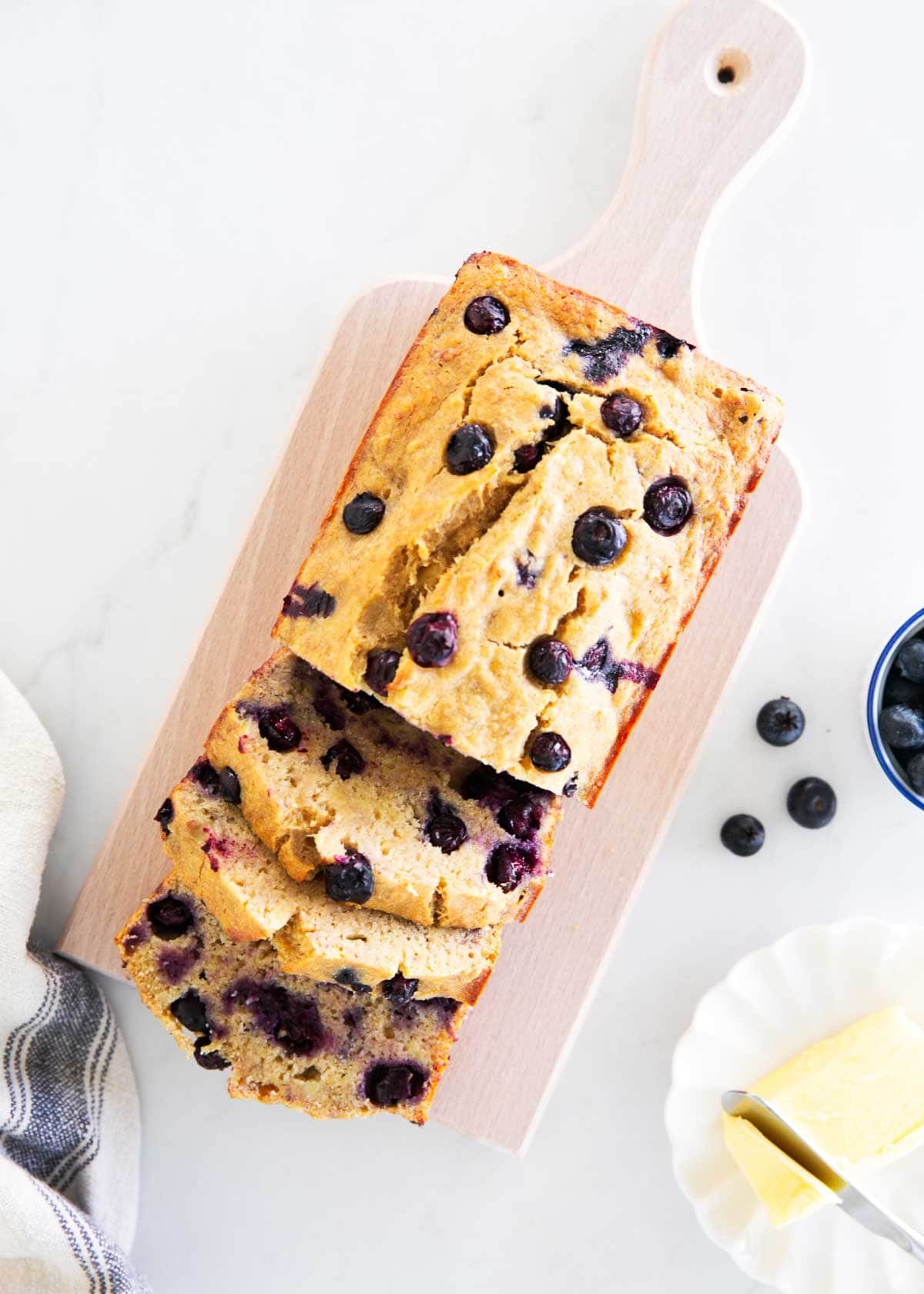 Bake up a moist and healthy blueberry banana bread in just over an hour using coconut oil, whole wheat flour, maple syrup, and loads of sweet bananas and juicy blueberries! Overhead view of sliced blueberry banana bread on a wooden serving board.