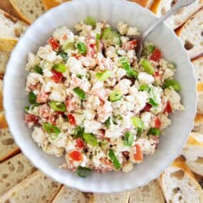 Overhead view of feta dip topped with green onions and tomatoes, surrounded by sliced bread on a wooden board.