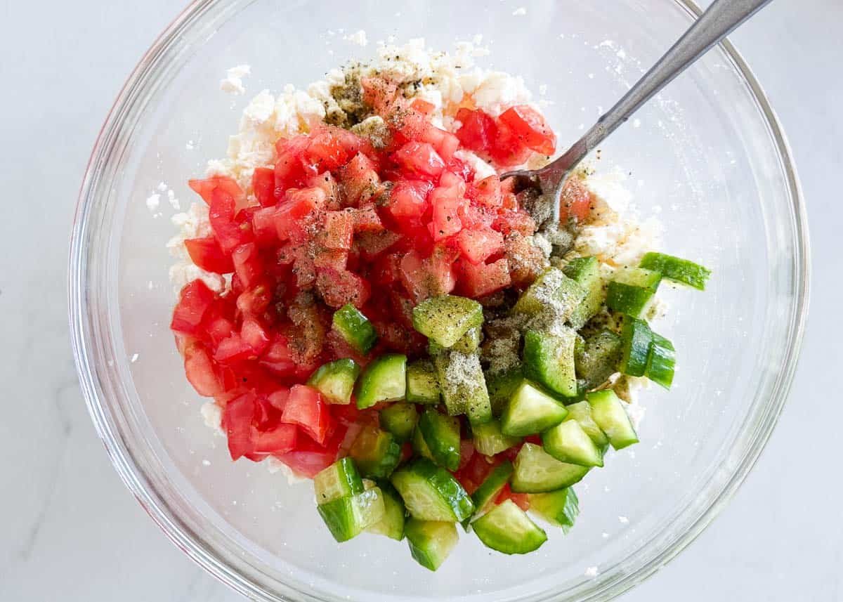 This feta dip comes together in 5 minutes with crumbled feta cheese, fresh vegetables, olive oil, and Greek seasoning. Serve it with crusty bread for an effortless appetizer. Feta dip ingredients being mixed in a bowl with crumbled feta, chopped tomatoes, cucumber, green onions, olive oil, and Greek seasoning.
