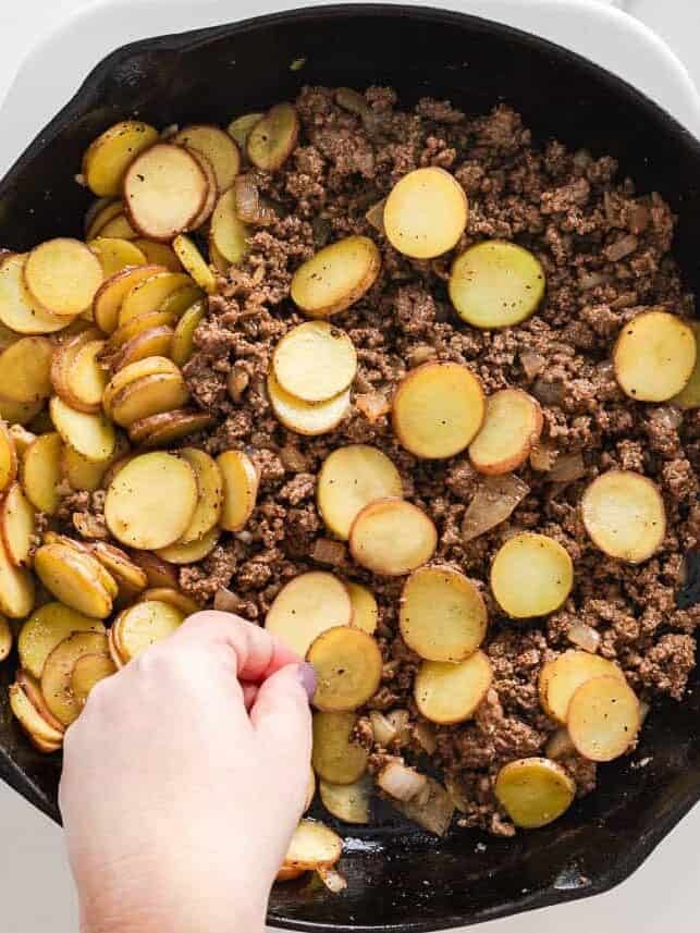 Seasoning ground beef and sliced potatoes together in a cast iron skillet.