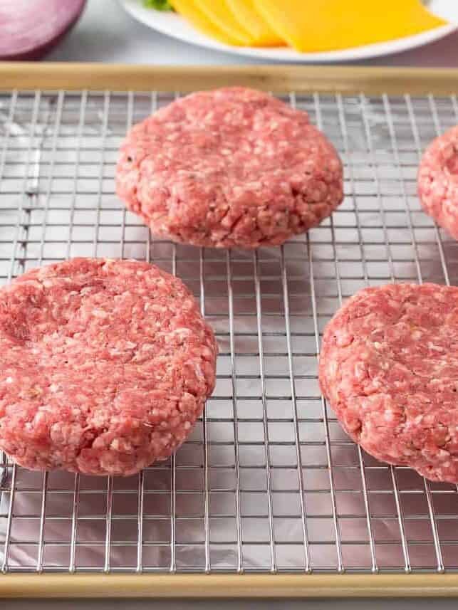 Raw hamburger patties arranged on a wire rack over a baking sheet, ready for cooking hamburgers in the oven.