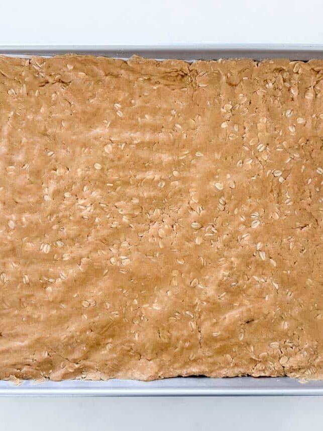 These lunch lady peanut butter bars are sweet and salty with a chewy peanut butter oatmeal cookie base, chocolate frosting on top and more peanut butter in between! Lunch lady peanut butter bars spread into a sheet pan before adding frosting.