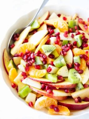 Overhead view of winter fruit salad with sliced apples, diced kiwi, peeled clementines, and citrus dressing in a white bowl.