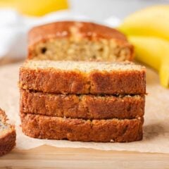 Three slices of sour cream banana bread stacked on parchment with bananas nearby.