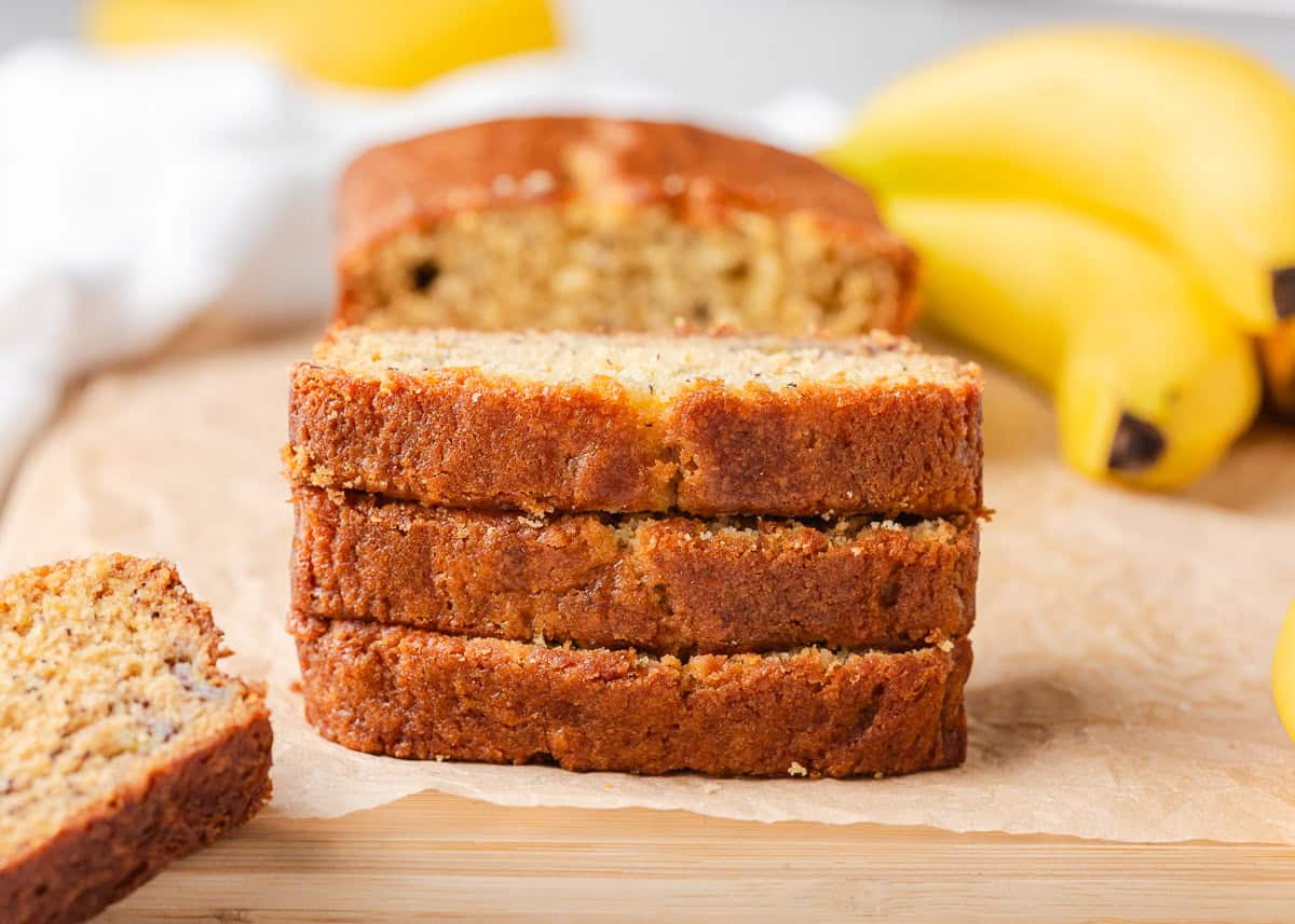 Three slices of sour cream banana bread stacked on parchment with bananas nearby.