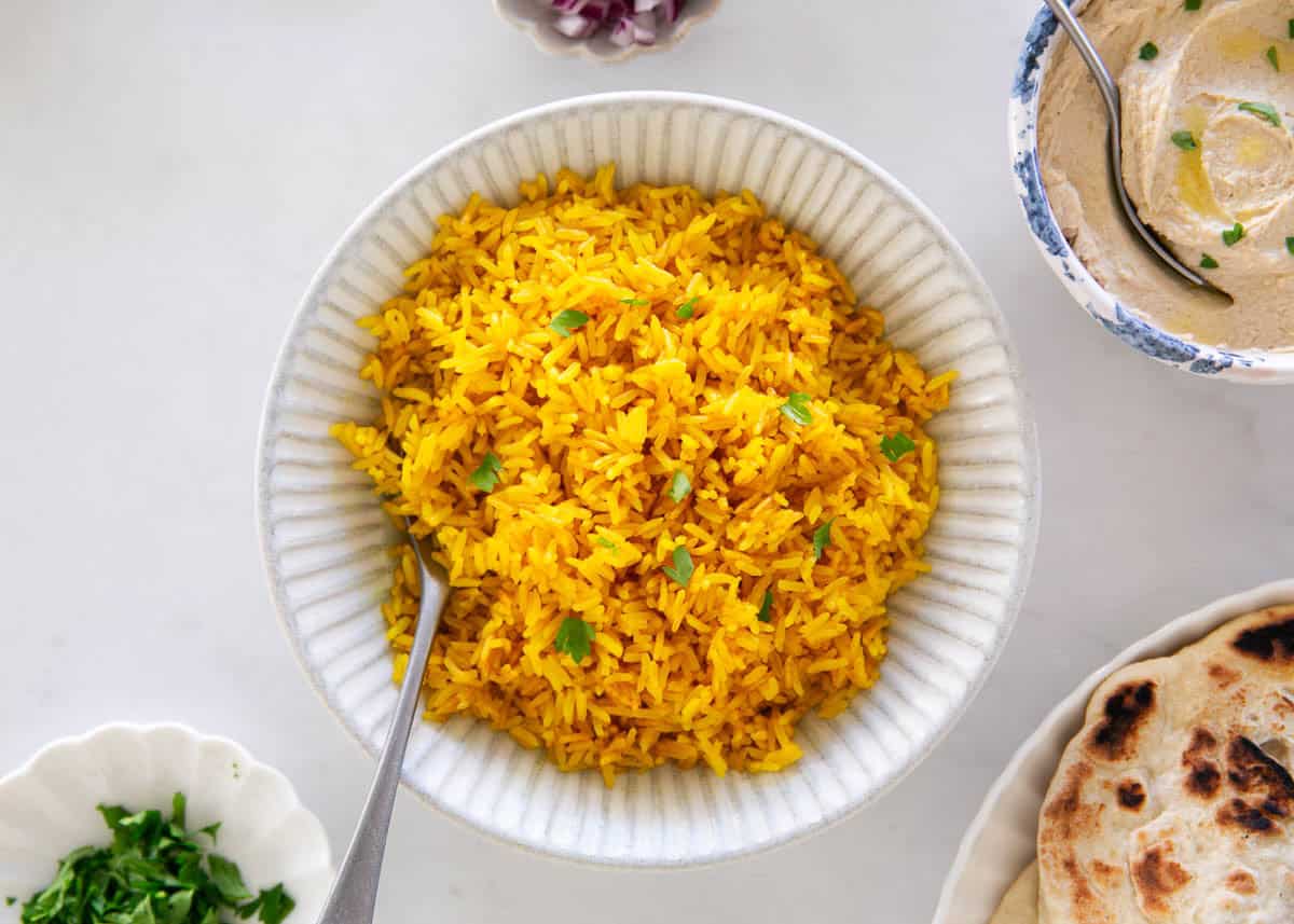 This easy yellow rice is cooked on the stovetop with chicken broth and turmeric for a fluffy, flavorful side dish recipe that elevates any meal and is ready in only 25 minutes! Overhead view of yellow rice served in a bowl with flatbread and hummus on the side.