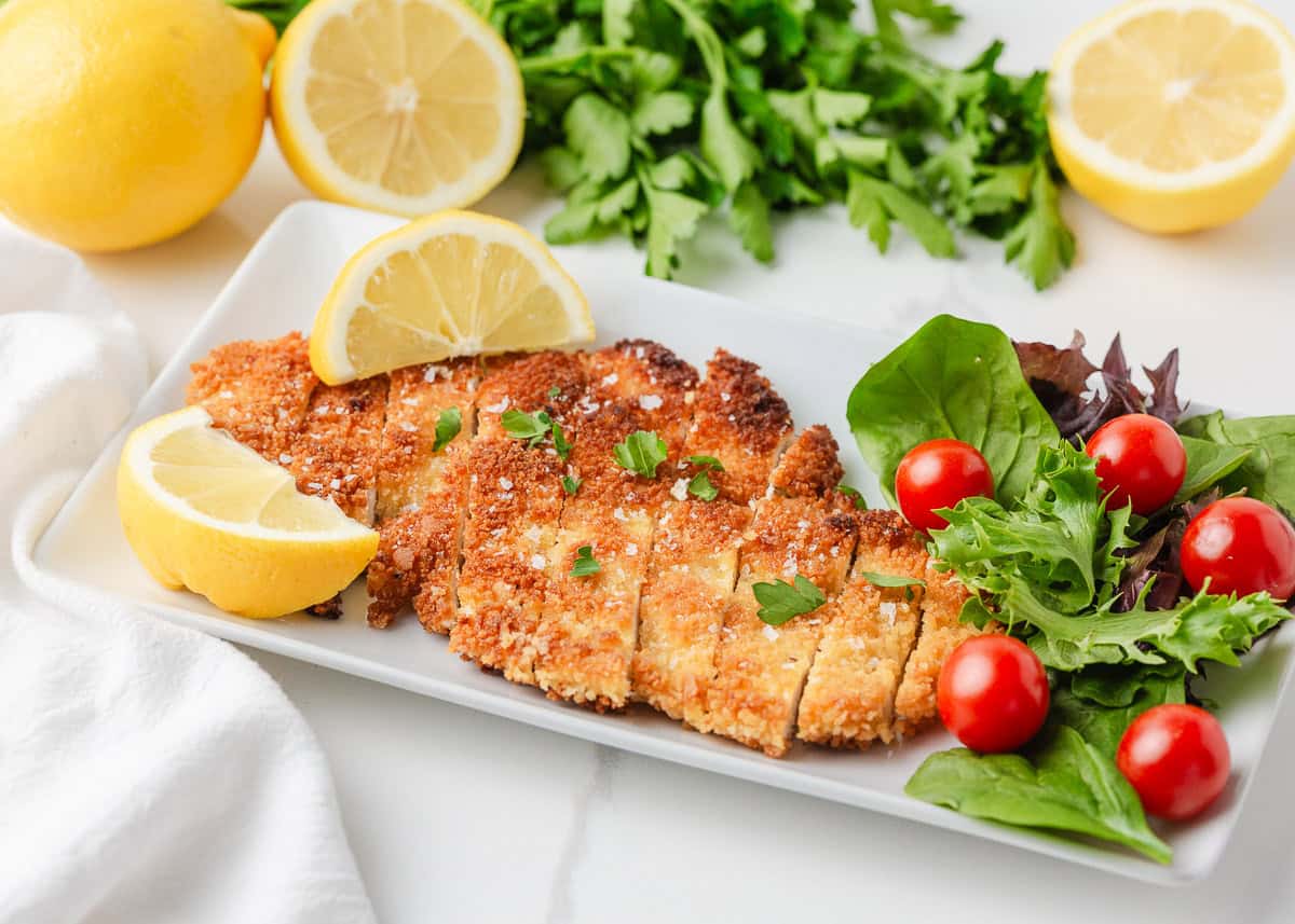 This chicken schnitzel is coated in seasoned breadcrumbs and pan fried until perfectly crispy, then finished with a squeeze of lemon. Thin, juicy, and ready in 30 minutes! Sliced chicken schnitzel showing a juicy interior and crunchy coating, served with a simple green salad and cherry tomatoes.